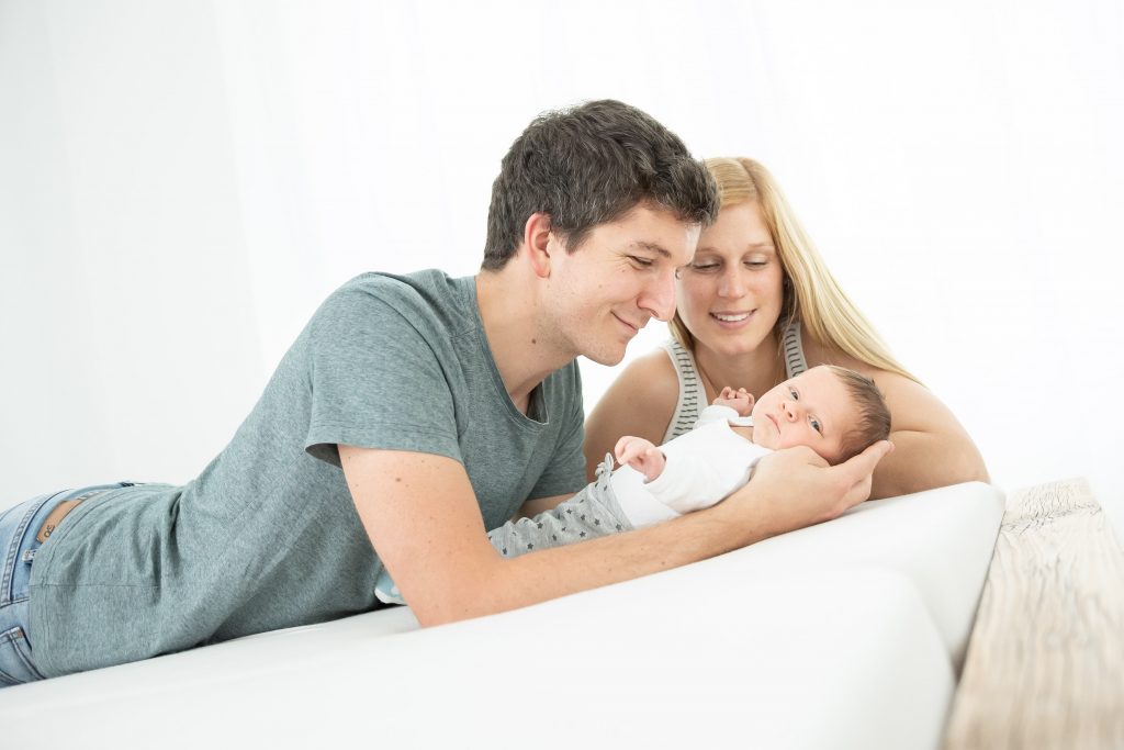 Ein Familienfoto zeigt ein Baby mit Mama und Papa. Die Eltern und das Kind scheinen zuhause fotografiert zu werden auf ihrem eigenem Bett in Ihrem Schlafzimmer. es ist ein sehr helles und freundliches Familienfoto mit viel Licht und glücklichen Eltern. Das Baby schaut entspannt in die Kamera und liegt in den Armen des Vaters.