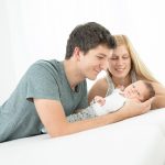 Ein Familienfoto zeigt ein Baby mit Mama und Papa. Die Eltern und das Kind scheinen zuhause fotografiert zu werden auf ihrem eigenem Bett in Ihrem Schlafzimmer. es ist ein sehr helles und freundliches Familienfoto mit viel Licht und glücklichen Eltern. Das Baby schaut entspannt in die Kamera und liegt in den Armen des Vaters.