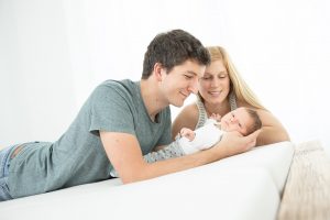Ein Familienfoto zeigt ein Baby mit Mama und Papa. Die Eltern und das Kind scheinen zuhause fotografiert zu werden auf ihrem eigenem Bett in Ihrem Schlafzimmer. es ist ein sehr helles und freundliches Familienfoto mit viel Licht und glücklichen Eltern. Das Baby schaut entspannt in die Kamera und liegt in den Armen des Vaters.