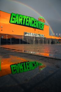 Der Globus Baumarkt in Herbolzheim ist für eine Imagewerbung und andere Werbung fotografiert. Es ist ein Architekturfoto von einem Fotografen in der Region und Umgebung von Herbolzheim, Ettenheim und Ortenau. Es ist ein Regenbogen zu sehen und dunkler bewölkter Himmel was sehr dynamisch und aufregend wirkt. Die Perspektive ist so gewählt, dass sich in einer Pfütze das Logo spiegelt. Das Gartencenter Globus Logo.