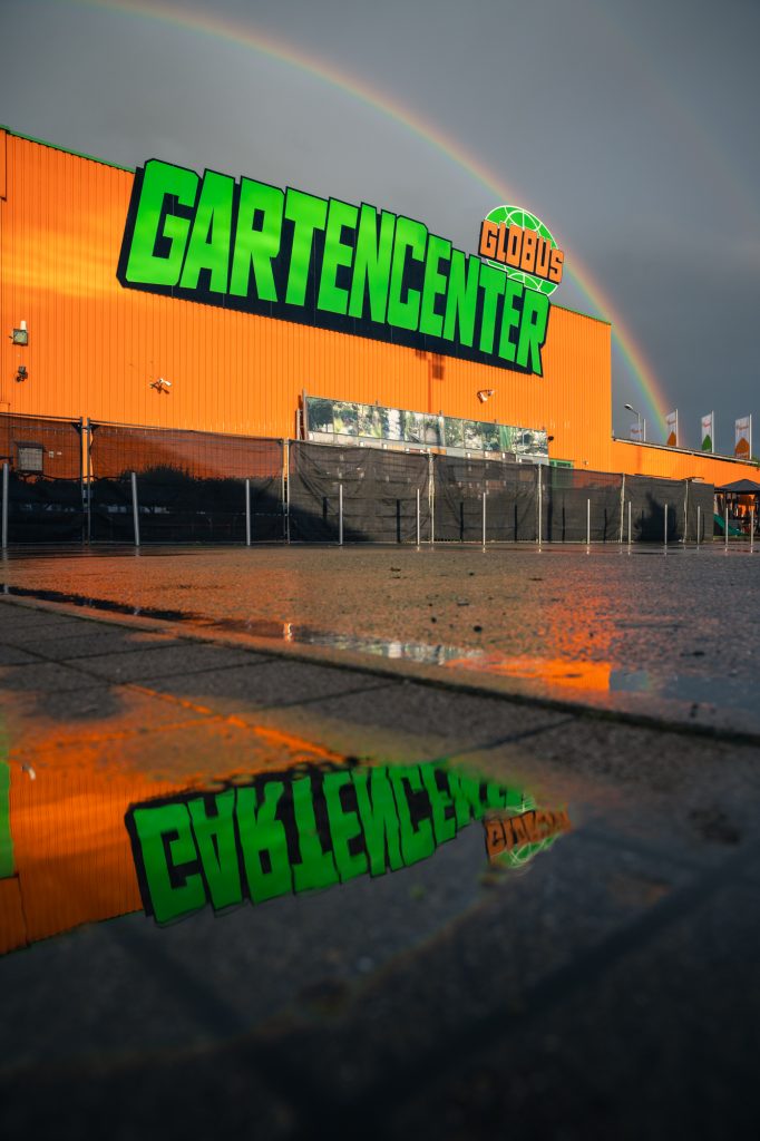 Der Globus Baumarkt in Herbolzheim ist für eine Imagewerbung und andere Werbung fotografiert. Es ist ein Architekturfoto von einem Fotografen in der Region und Umgebung von Herbolzheim, Ettenheim und Ortenau. Es ist ein Regenbogen zu sehen und dunkler bewölkter Himmel was sehr dynamisch und aufregend wirkt. Die Perspektive ist so gewählt, dass sich in einer Pfütze das Logo spiegelt. Das Gartencenter Globus Logo.